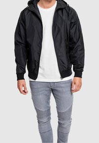 Svart huvjacka i bomberstil gjord av glänsande material, med dragkedja framtill, ribbade mudd och sidofickor. Matchad med ljusgrå slim-fit jeans.