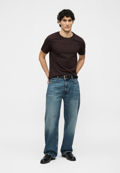 Νέος άνδρας με σγουρά μαλλιά όρθιος, φοράει σκούρο καφέ μπλουζάκι, φαρδιά μπλε τζιν με μαύρη ζώνη και μαύρα παπούτσια σε μονόχρωμο φόντο.
