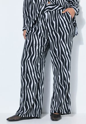 Kobieta w czarno-białych szerokich spodniach w paski zebry i dopasowanej bluzce, z jedną ręką w kieszeni i czarnych plecionych balerinach.