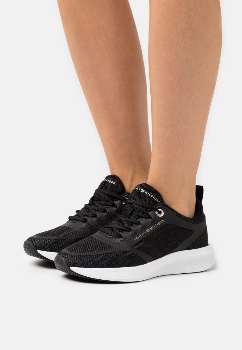 Baskets à lacets noires Tommy Hilfiger avec semelles blanches portées pieds nus sur un fond blanc uni.