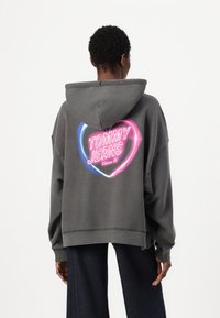 Haut à capuche surdimensionné gris avec un grand motif rose et bleu au dos affichant "TOMMY JEANS Depuis 85." Il est doté d'un capuchon avec cordon de serrage et de poignets côtelés.