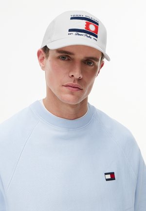 Vyras dėvi baltą Tommy Hilfiger kepuraitę su raudonu ir mėlynu logotipu bei šviesiai mėlyną megztinį su mažu Tommy Hilfiger vėliavos emblematu ant krūtinės.