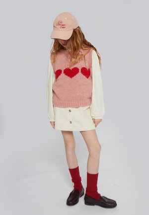 Chaleco rosa de punto con patrón de corazón rojo, camiseta de manga larga color crema, falda de pana color crema, calcetines rojos acanalados y zapatos tipo mocasín negros.