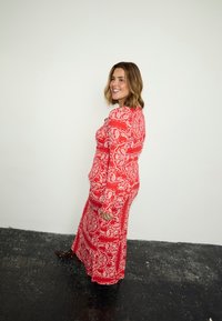 Γυναίκα που χαμογελά και περπατά, φορώντας ένα μακρύ κόκκινο φόρεμα με μοτίβο παϊέτας και καφέ μπότες με τακούνι, μπροστά σε απλή λευκή τοιχοποιία.