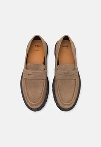 Mocassins en daim marron avec un design de mocassin, ornés d'une couture, semelle en caoutchouc et intérieur beige avec une marque sur la semelle intérieure.