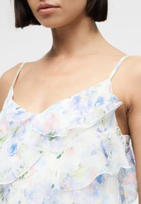 Γυναίκα που φοράει ένα διάφανο λευκό φόρεμα με λουλούδια, με παστέλ μπλε, ροζ και πράσινα στρώματα με βολάν και λεπτούς ιμάντες στους ώμους.