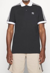 Man i svart Adidas polo med vit krage och ränder, samt beiga Adidas shorts med svarta ränder och logotyp.