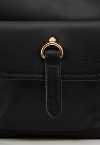 Dettaglio di una borsa a mano nera in pelle con anello e tracolla in metallo dorato. Superficie opaca con cuciture visibili lungo i bordi.