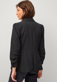 Zwarte blazer met een getailleerde pasvorm, gestructureerde schouders en gathered mouwen. Een gladde textuur en strakke lijnen voor een verzorgde uitstraling.