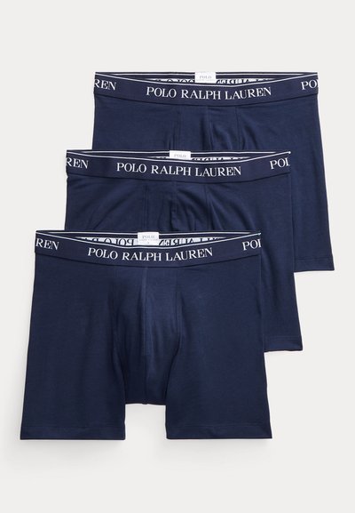 Trys tamsiai mėlynos Polo Ralph Lauren šortų apatiniai su juodu juosmeniu, kuriame yra baltos spalvos prekės ženklo užrašai, sudėti vienas ant kito.