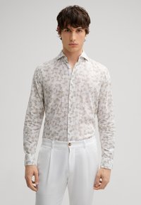 Weißes Langarmhemd mit hellbraunen Blumenmustern, mit Knopfleiste am Kragen und glatter Textur. Kombiniert mit weißen Hosen, die einen Knopf haben.