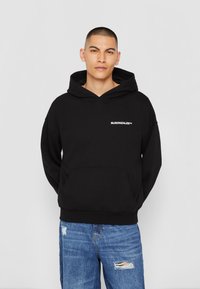 Haut à capuche noir avec une poche avant, arborant le texte blanc "MJGONZALES™" sur la poitrine, porté avec un jean déchiré bleu clair.