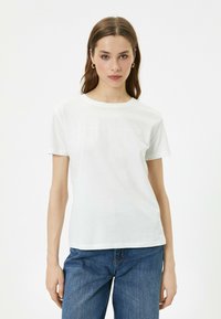 Weißes Baumwoll-T-Shirt mit kurzen Ärmeln, Rundhalsausschnitt und subtilen Textdesign in hellen Farben auf der Vorderseite. Kombiniert mit blauen Denim-Jeans.