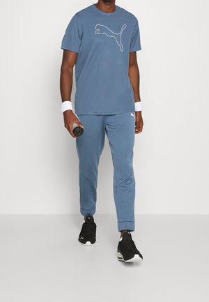 Blue athletic shirt with white Puma logo, paired with matching blue pants and black sneakers. Sweatbands on wrists, holding a water bottle.