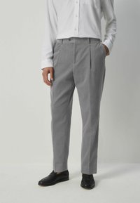 Pantalones gris ajustados con una textura sutil, frente plisado y bolsillos, combinados con una camisa blanca de manga larga y mocasines negros.
