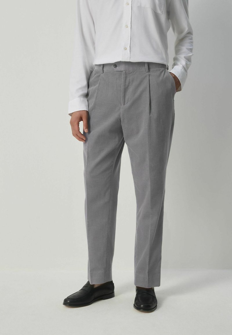 Pantalones gris ajustados con una textura sutil, frente plisado y bolsillos, combinados con una camisa blanca de manga larga y mocasines negros.