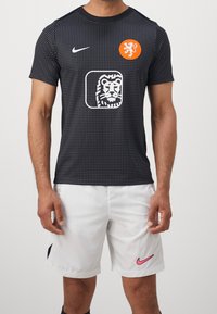Svart sporttröja med prickigt mönster, med en orange rund logotyp och en lejon- grafik. Matcher med vita shorts och rosa Nike-logotyp.