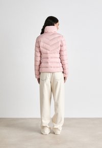 Ροζ καπιτονέ μπουφάν με ψηλό γιακά, με σχέδιο διαγωνίου καπιτονέ. Συνδυάζεται με φαρδιά, κρεμ παντελόνια.