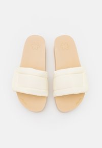 Cream-colored slide sandals with padded straps, smooth tan footbed, and a flat sole; minimalistic design with embossed logo detail.