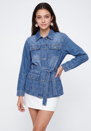 Jonge vrouw draagt een blauwe denimjas met ceintuur, voorzakken en knoopdetails, gecombineerd met een witte rok en oorbellen in bloemvorm.