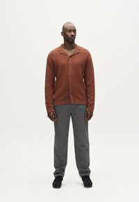 Maglione ribbed color ruggine con colletto a scollo, chiusura con bottoni, indossato con pantaloni grigi e scarpe nere. Design semplice e strutturato.