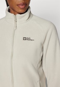 Giacca in pile crema con colletto alto e zip completa. Presenta un piccolo logo ricamato sul lato sinistro del petto. Texture morbida e design casual.