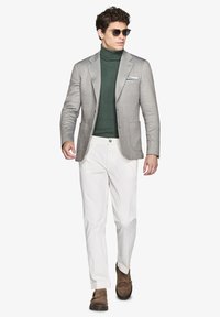 Lys grå blazer, mørkegrøn ribbet turtleneck, hvide bukser og brune ruskinds sko. Brillerne er mørke og runde.