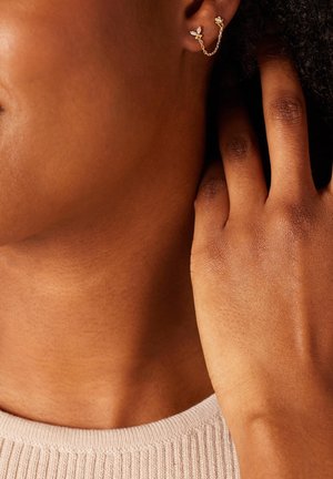 Les boucles d'oreilles en chaîne dorée présentent des détails floraux et un maillon délicat. La texture est lisse, complétant le teint de la peau et le fond en tissu tricoté.