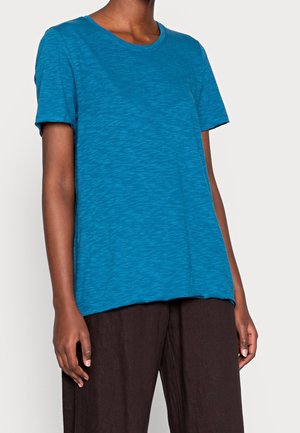 Person trägt ein kurzärmliges türkisblaues T-Shirt und lockere schwarze Hose, dargestellt vom Hals bis zur Mitte des Oberschenkels vor einfachem Hintergrund.