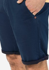 Navy blue shorts made of cotton, featuring rolled cuffs, side pockets, and a small branded button detail. Smooth texture, casual design.