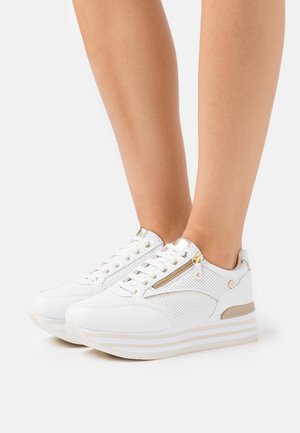 Baskets montantes blanches avec fermeture éclair dorée et accents dorés portées sur des jambes nues sur un fond blanc uni.