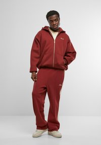 Felpa rossa con zip, tasche frontali e polsini a costine, abbinata a pantaloni della tuta con righe laterali color sabbia e logo ricamato.