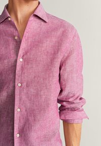 Lichtroze linnen shirt met een subtiele textuur, knoopsluiting en opgerolde mouwen. Voorzien van witte knopen langs de voorkant.