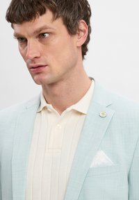 Lichtblauwe blazer in unstructured stof, gecombineerd met een crèmekleurig overhemd met kraag. Bevat een witte pochet en een bloemvormige reversspeld.