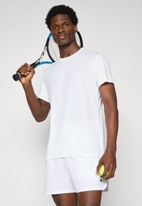 Hvit idrettsskjorte med kortermer og rund hals, kombinert med hvite shortser; holder en tenniskølle og en grønn tennisball.