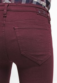 Närbild av en person som bär mörkröda jeans med synlig sömnad på bakfickorna och en mönstrad skjorta instoppad.