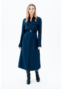 Eine Frau trägt ein langärmliges, marineblaues, mit Gürtel versehenes Hemdkleid und schwarze kniehohe Stiefel und steht vor einem weißen Hintergrund.