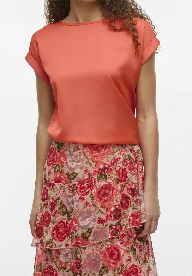 Femme portant un chemisier corail à manches courtes associé à une jupe fleurie à volants avec des roses rouges et roses et des feuilles vertes.