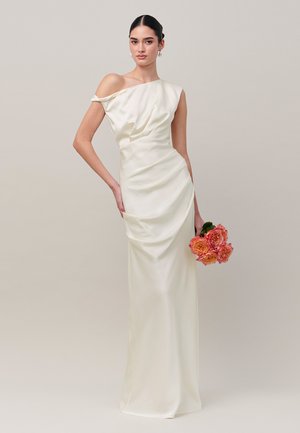 Frau in einem weißen Satin-Ein-Schulter-Kleid, die einen kleinen Strauß aus orangefarbenen und rosa Rosen vor einem schlichten neutralen Hintergrund hält.