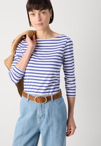Stripet langermet topp i hvit og blå, kombinert med lyse denim bukser og et brunt belte. Modellen holder en beige jakke på en avslappet måte.