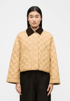 Femme aux cheveux noirs raides portant une veste matelassée beige avec un col oversize marron foncé et un pantalon noir, debout devant un fond uni.