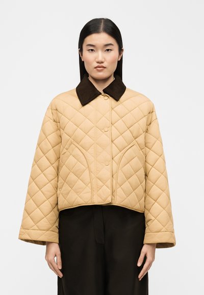 Femme aux cheveux noirs raides portant une veste matelassée beige avec un col oversize marron foncé et un pantalon noir, debout devant un fond uni.
