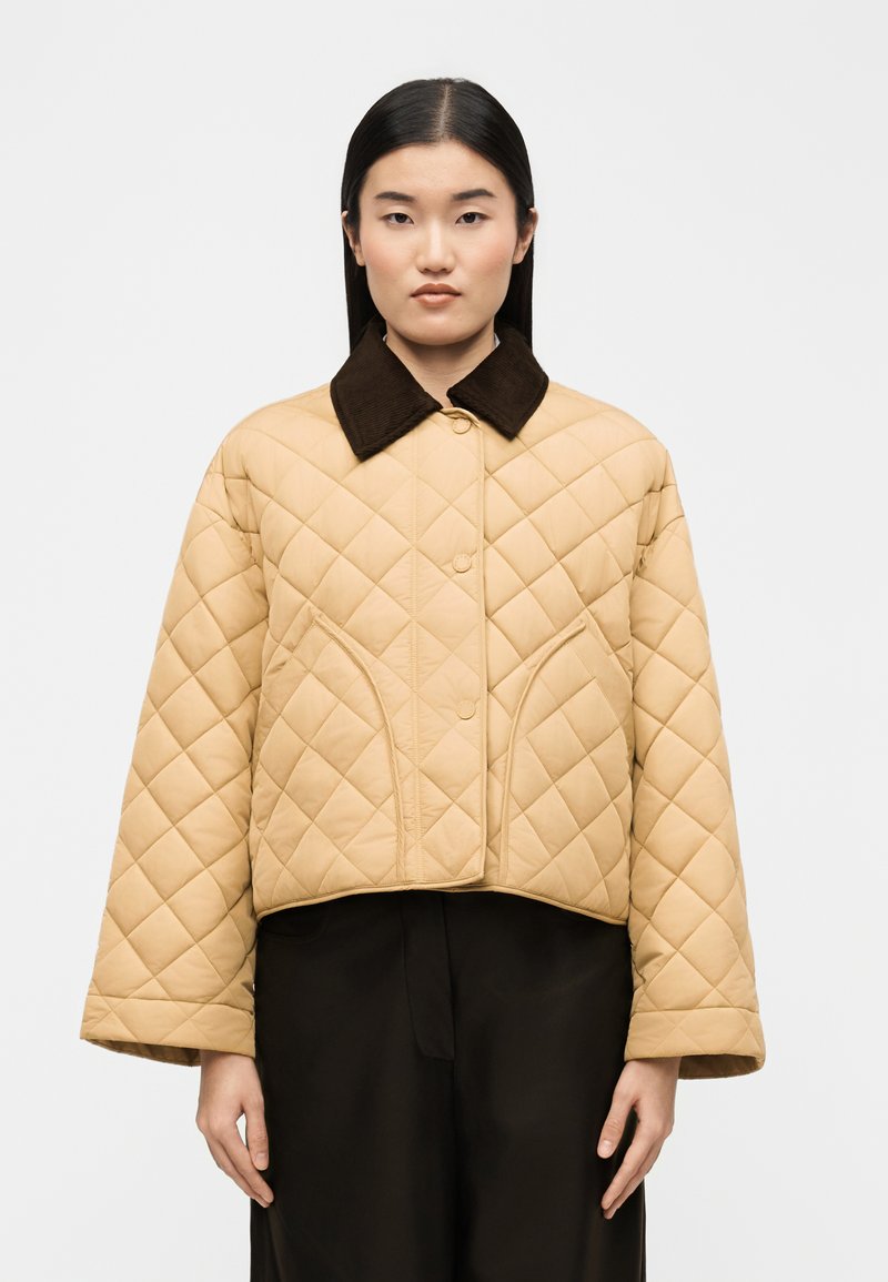 Γυναίκα με ίσια μαύρα μαλλιά φοράει μπεζ καπιτονέ μπουφάν με υπερμεγέθη σκούρο καφέ γιακά και μαύρο παντελόνι, στέκεται μπροστά σε απλό φόντο.