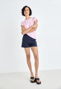 Randig rosa och vit kortärmad skjorta i kombination med marinblå shorts. Modellen bär svarta, tunga sandaler. Slät tyg, avslappnad passform.