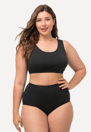 Smiling woman with long hair wearing a black sports bra and high-waisted black underwear, standing with one hand on her hip.