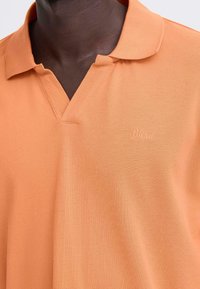 Polo à manches courtes en tissu texturé orange avec un col en V et un logo "Blend" brodé sur la poitrine. Coupe décontractée.