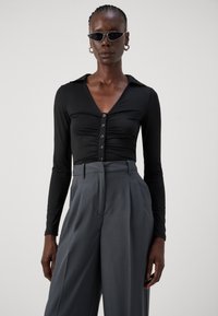Haut noir à manches longues avec un col en V, comportant des détails froncés et des boutons noirs, associé à un pantalon gris taille haute avec des plis.
