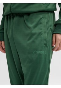 Pantalon de sport vert avec poche latérale et logo hummel sur la cuisse, porté avec une veste verte assortie.