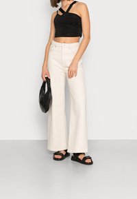 Svart kort t-shirt med korsande remmar, vita vida jeans och en svart texturerad handväska. Svarta sandaler med remmar framtill.