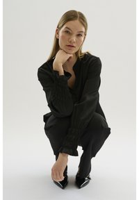 Schwarzes, seidiges Outfit mit gerafften Ärmeln, das locker geschnitten ist, und spitzen, schwarzen Absätzen. Das Model hockt vor einem neutralen Hintergrund.